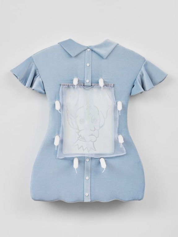 Blue dress with scalloped edges and a large embroidered image of a cartoon face in the center, mounted on a wall display.