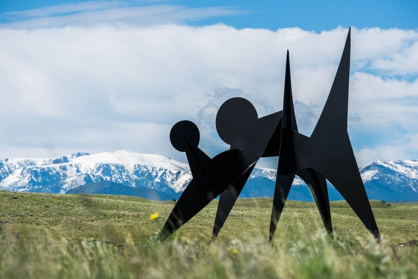 Abstract black metal sculpture with geometric shapes set against a backdrop of green fields and snow-capped mountains.