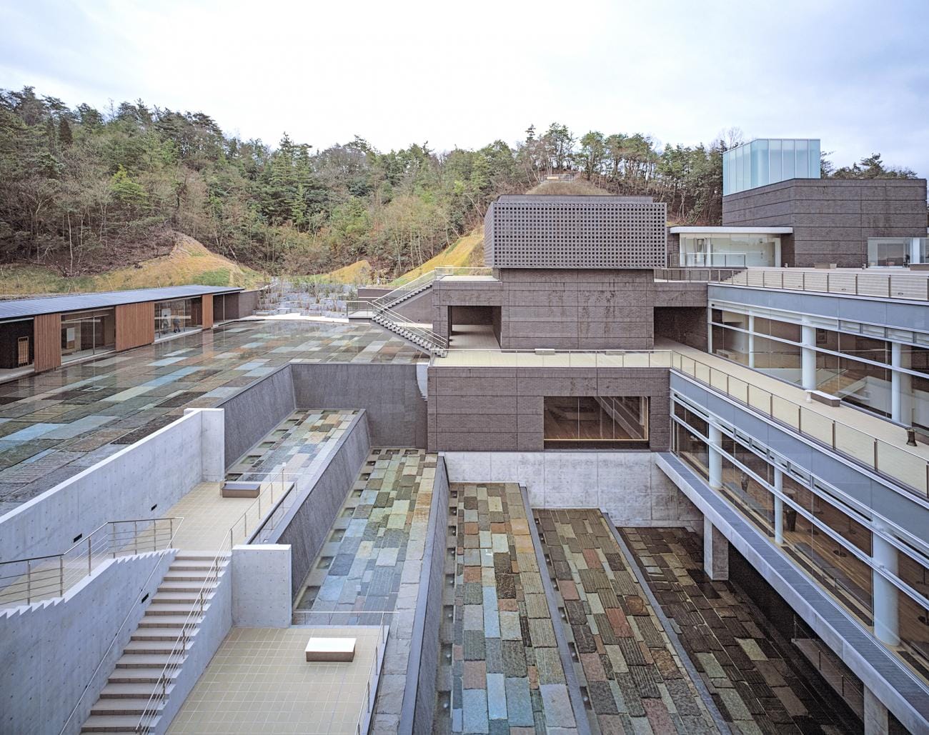 Modern building with textured stone pathways, surrounded by trees and featuring multi-level terraces with large windows.