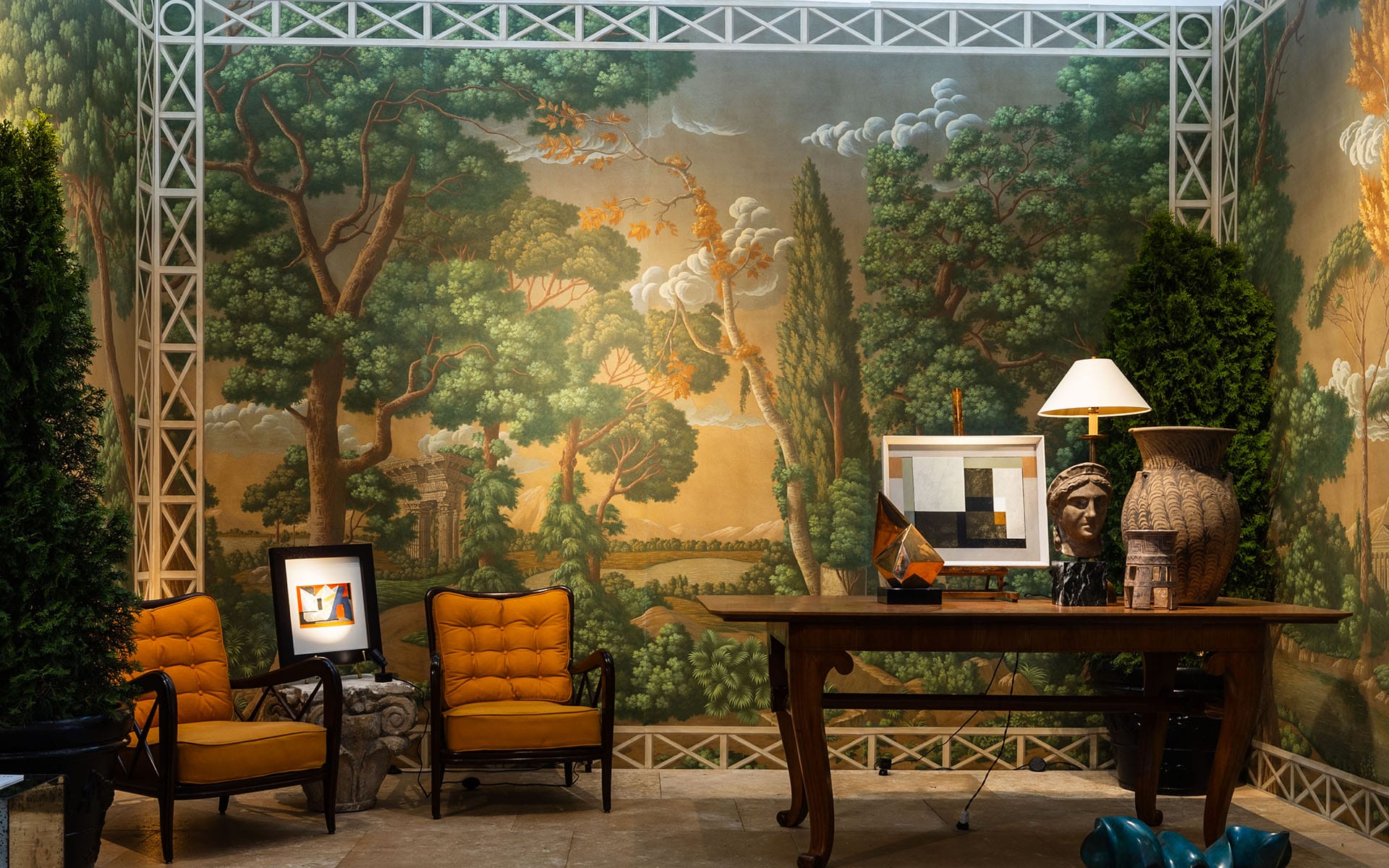 Elegant room with nature-themed mural, orange chairs, wooden table, artistic decor, and lush greenery.