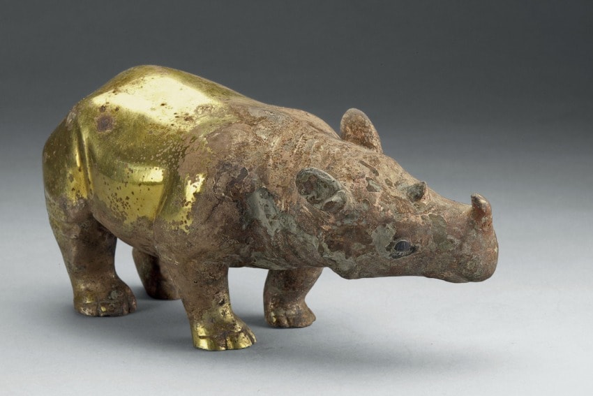 Ancient bronze and gold rhinoceros statue, with a weathered surface, displayed in a museum setting on a plain background.