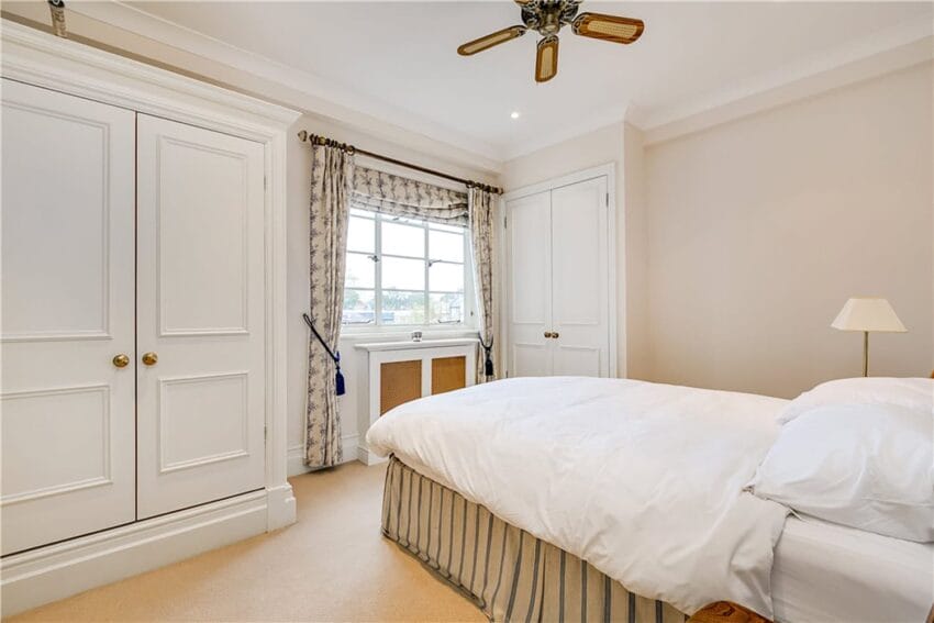 Bedroom with white bedding, ceiling fan, window with curtains, built-in wardrobes, and neutral walls.