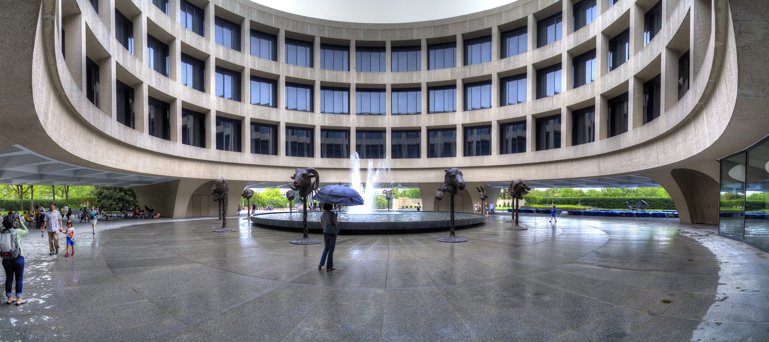 People walking near a fountain and elephant sculptures in a circular courtyard of a large modern building.