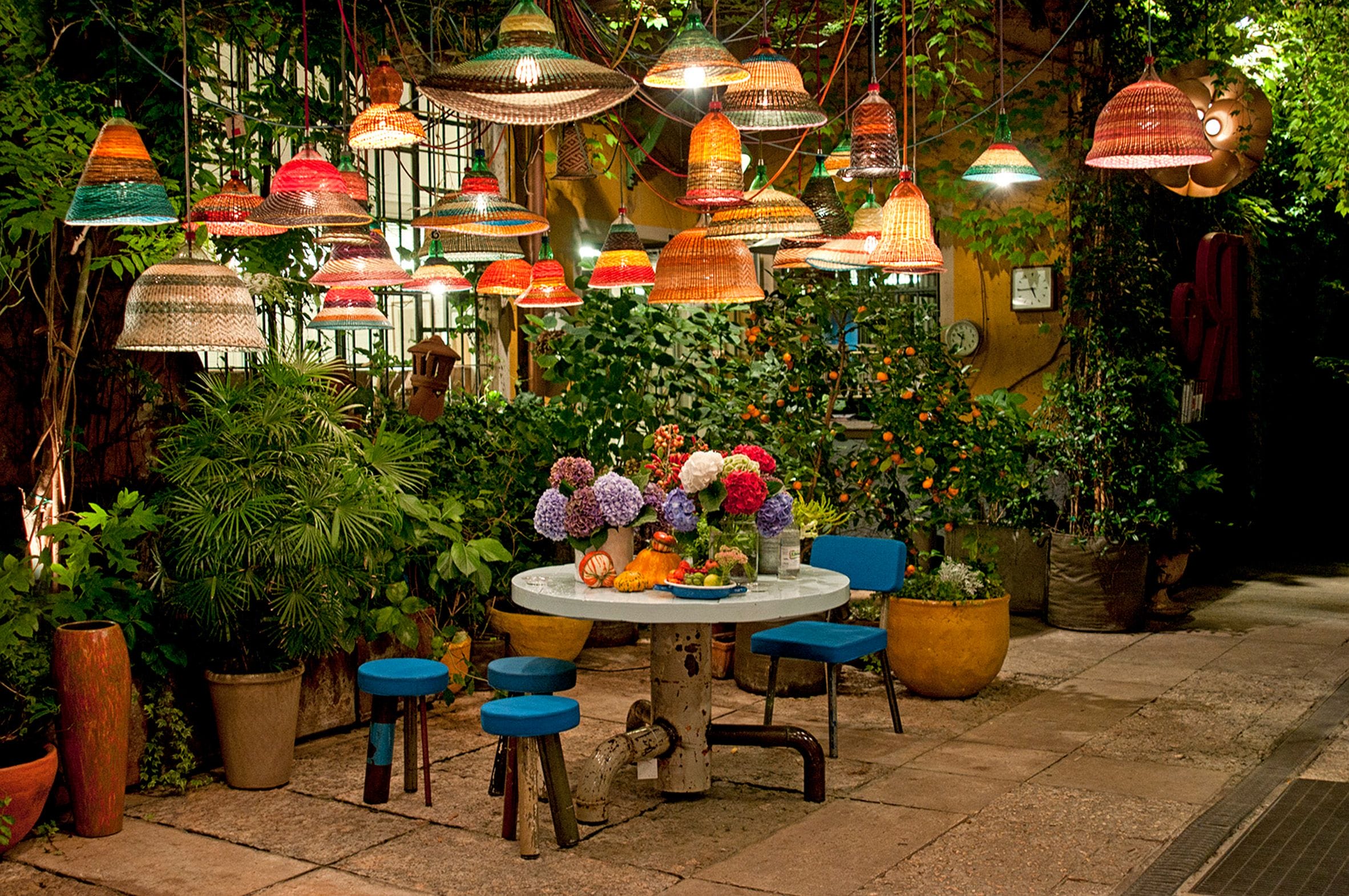 Outdoor patio with colorful hanging lights, a round table, blue stools, and vibrant flowers surrounded by lush greenery.