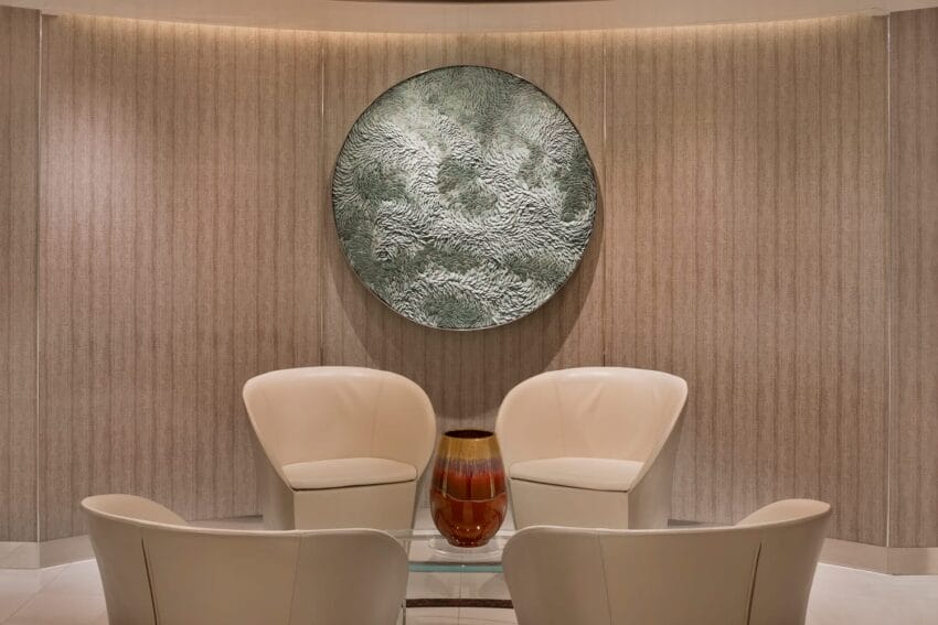 Modern lounge with four cream-colored chairs, glass table, decorative vase, and large textured circular wall art.