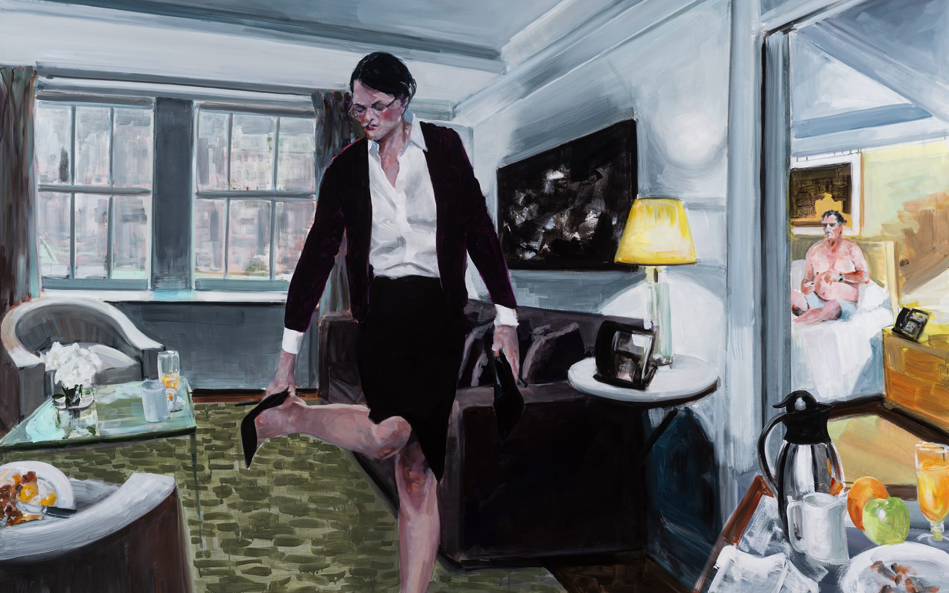 Person in a suit adjusts shoe in a modern living room, with another person visible in a distant room.