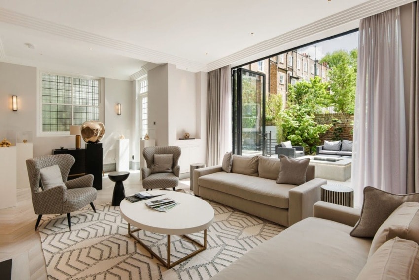 Modern living room with open patio doors to a garden, featuring cozy sofas, armchairs, a coffee table, and neutral decor.