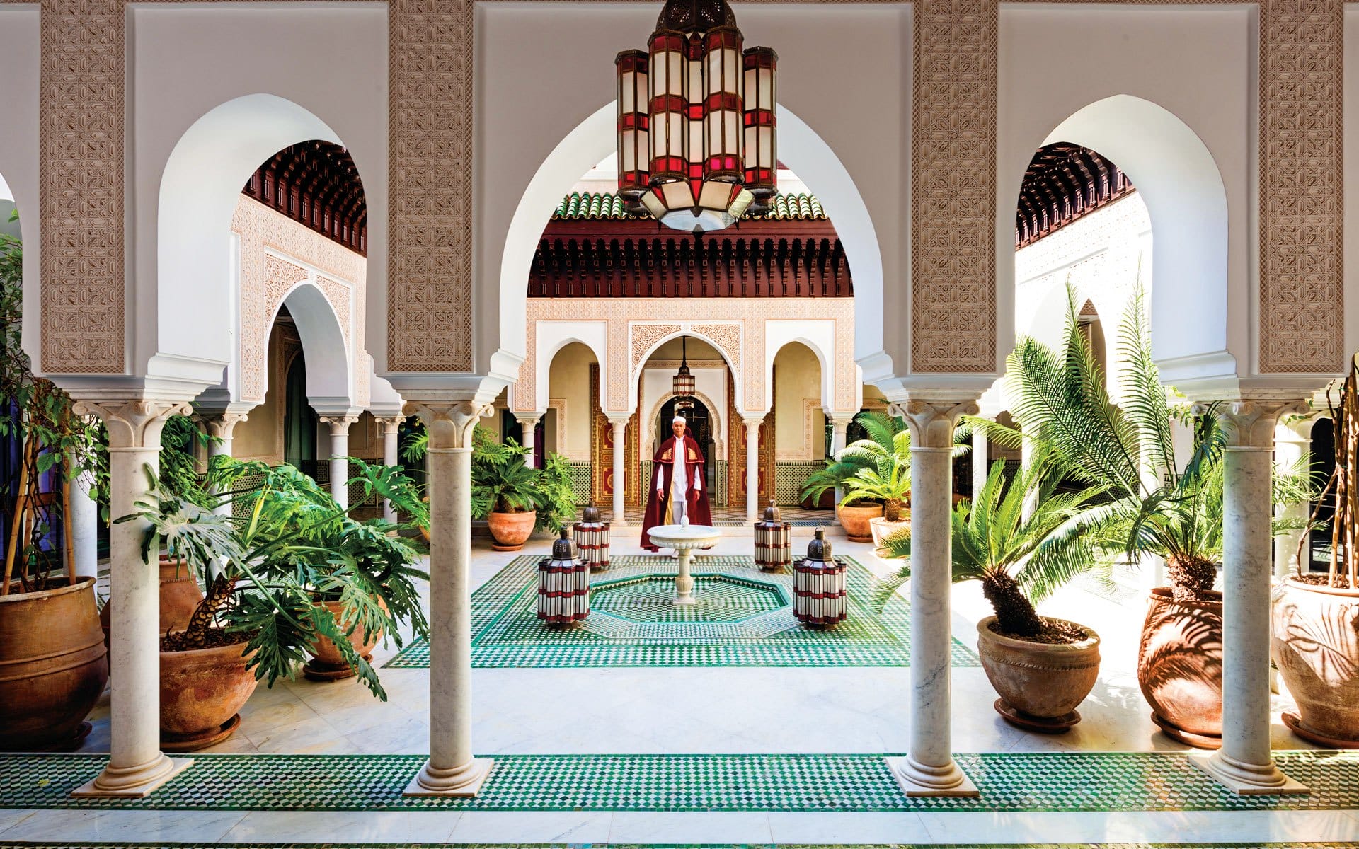 Elegant Moroccan courtyard with arches, lush plants, and intricate tile flooring, featuring a central fountain under a chandelier.