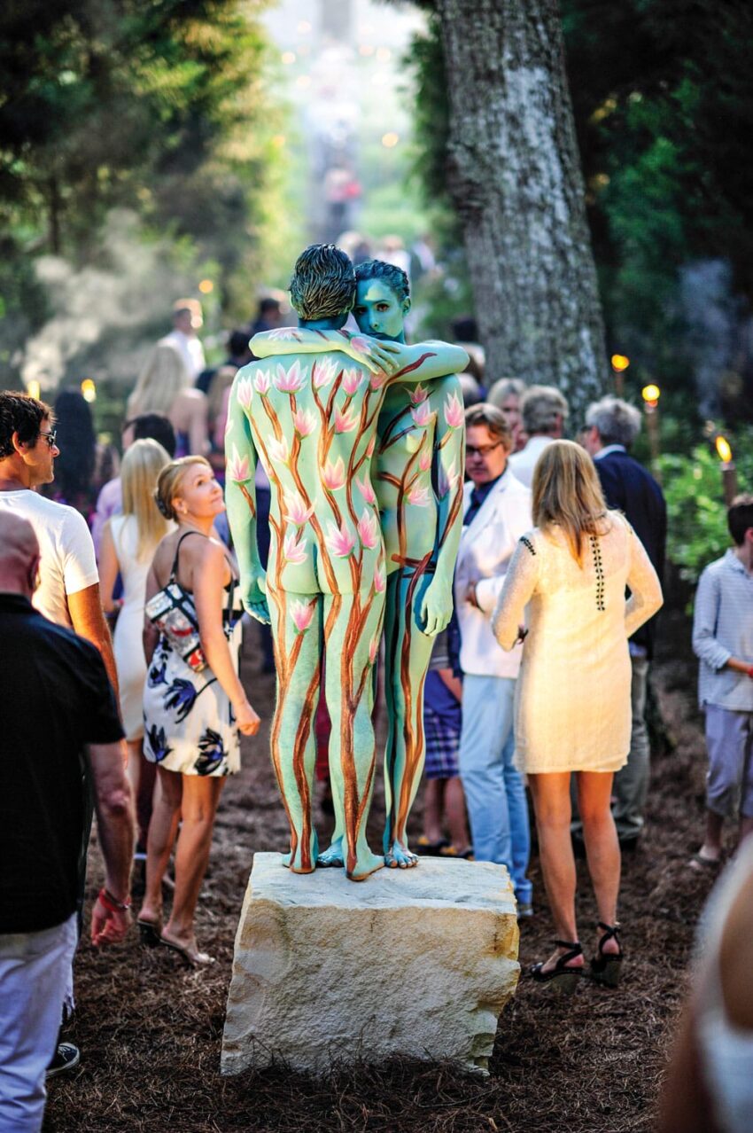 Two people painted with floral art stand on a pedestal in a wooded area surrounded by onlookers.