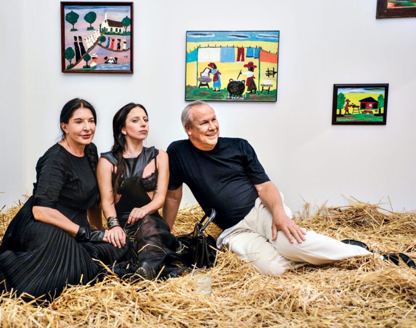 Three people sitting on straw with colorful paintings on the wall in the background.