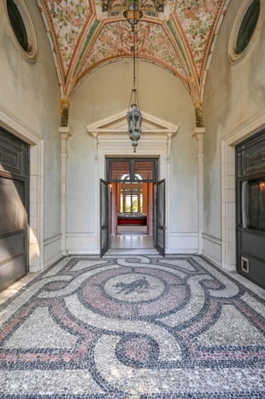 Elegant corridor with decorative mosaic floor, arched ceiling, and ornate chandelier, leading to an open doorway.