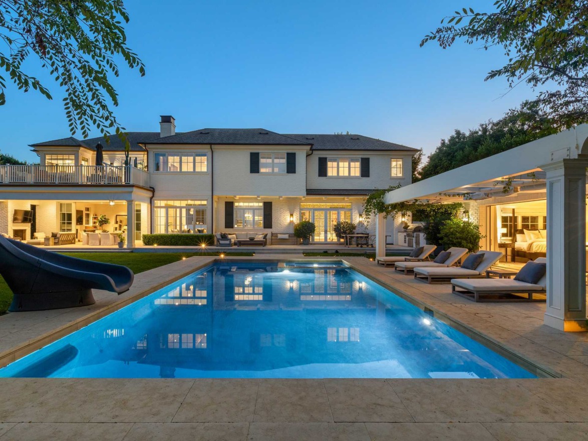 Luxurious house with a lit pool in the foreground, surrounded by trees and outdoor seating at dusk.
