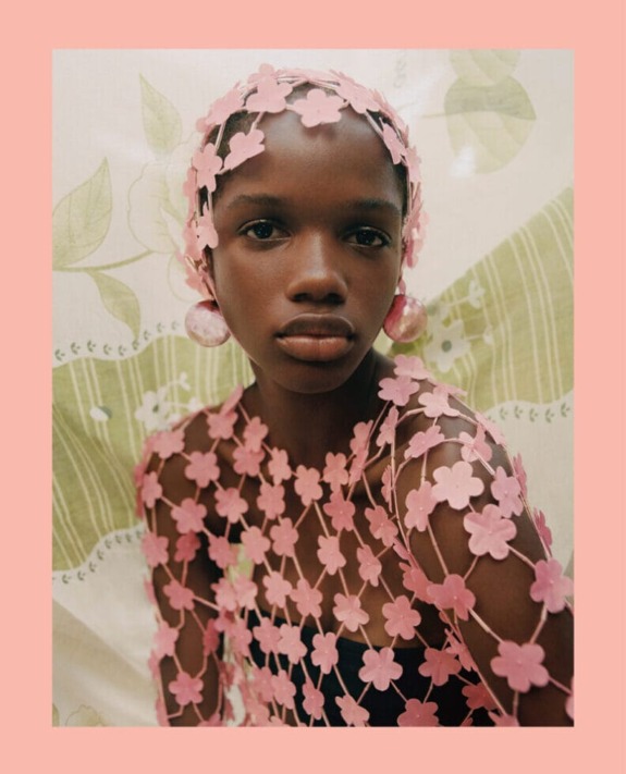 Model wearing a floral pink netted outfit with matching headpiece and large round earrings against a patterned background.