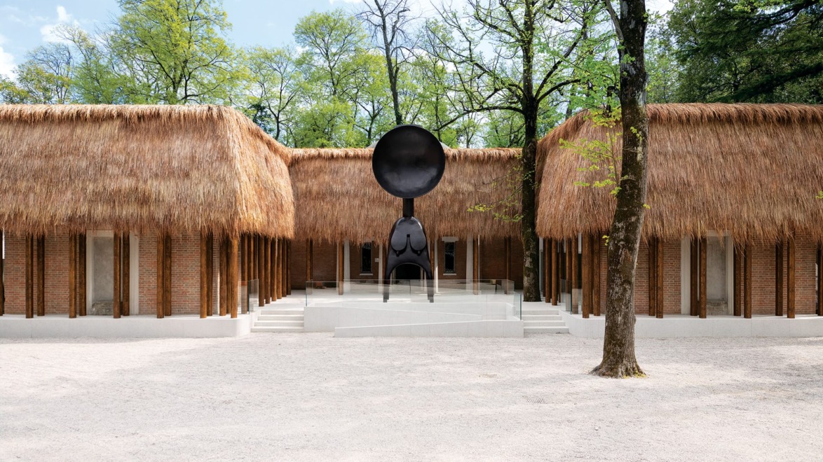 Thatched-roof buildings with a contemporary metal sculpture in a tree-lined courtyard on a sunny day.