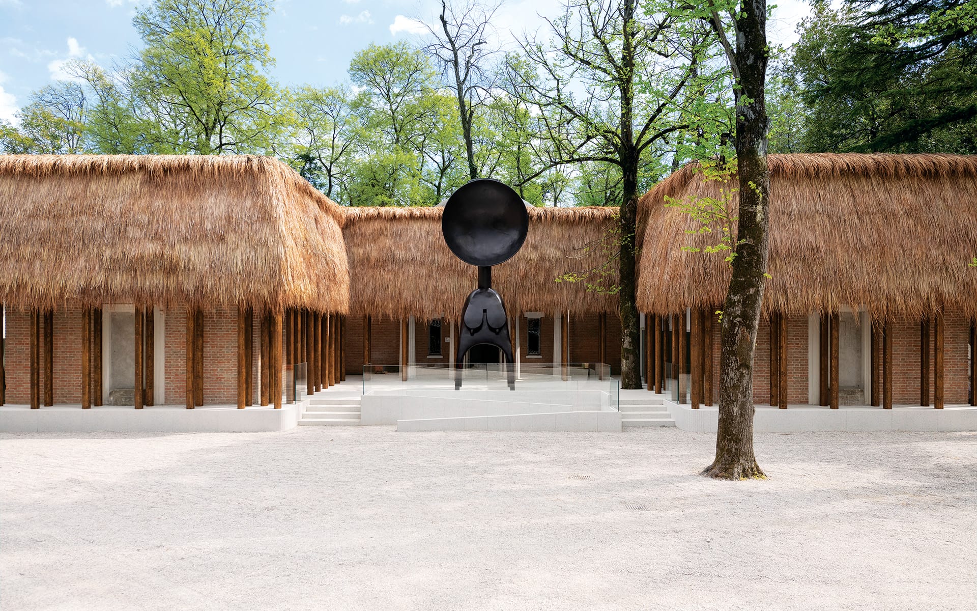Thatched-roof buildings with a contemporary metal sculpture in a tree-lined courtyard on a sunny day.