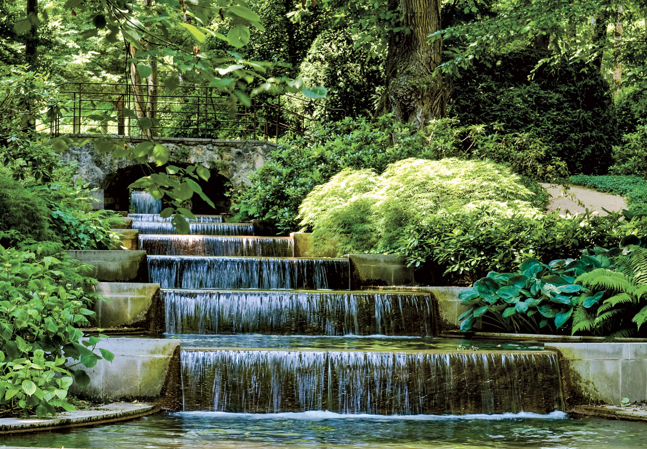 A serene garden waterfall cascading over stone steps surrounded by lush greenery and trees, with a small wooden bridge above.