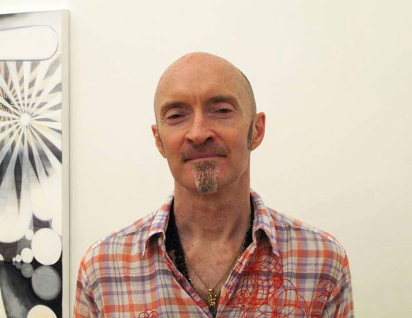 Person in a plaid shirt standing indoors with a painting in the background.