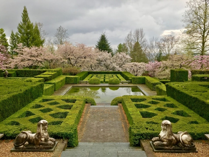 Formal garden with hedges, geometric paths, statues, and a central reflecting pool surrounded by blooming trees under cloudy sky.