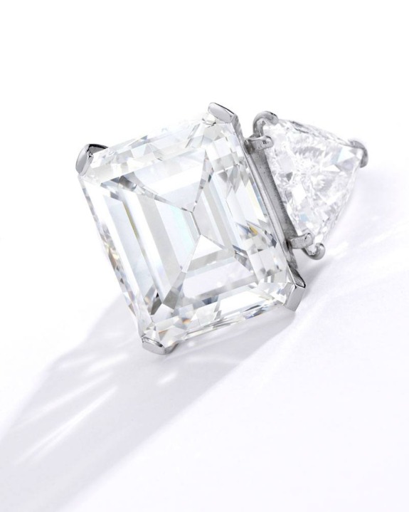 Emerald-cut diamond ring with a side triangular diamond on a reflective surface.