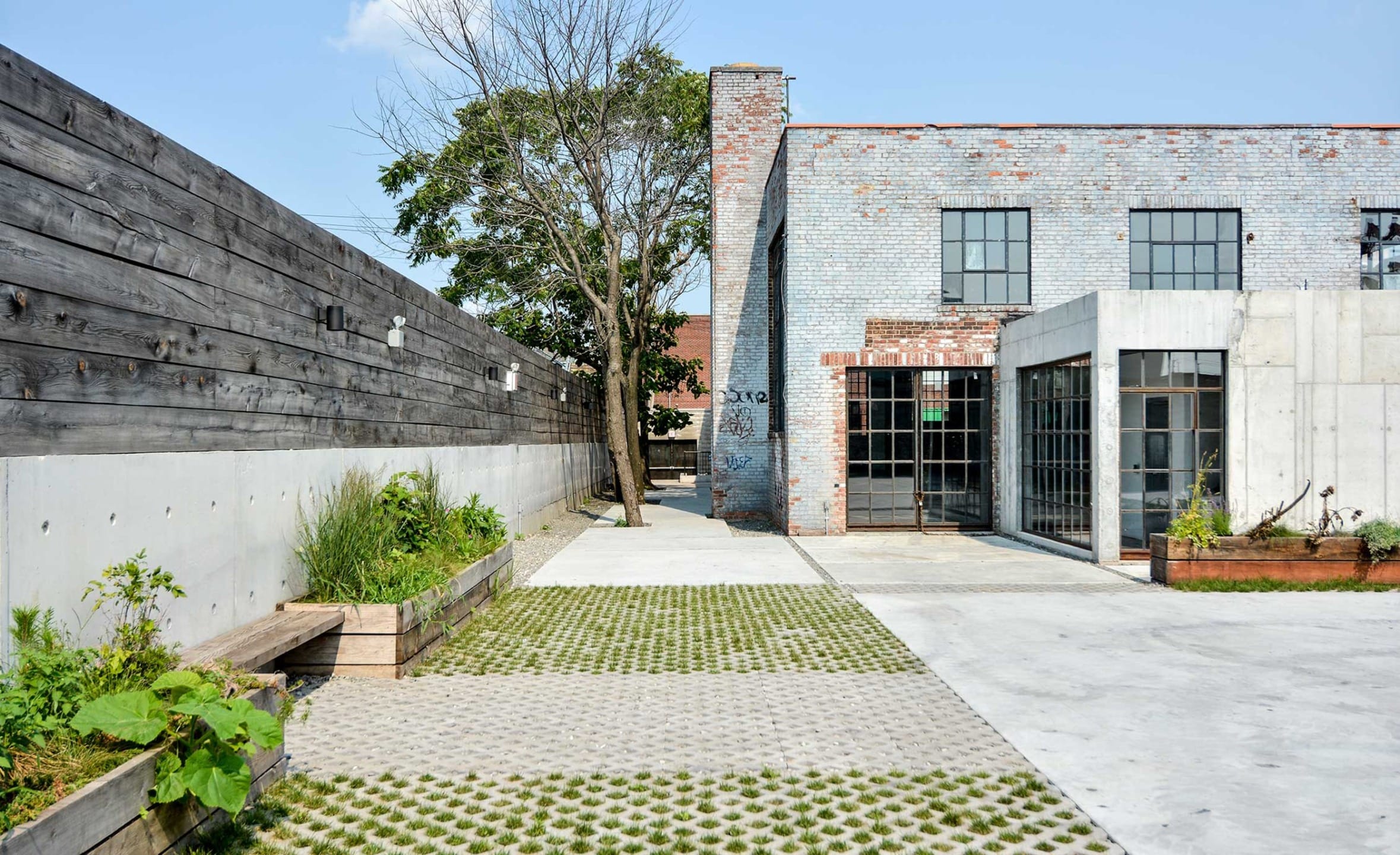 Modern industrial building with brick facade, large windows, and outdoor patio space featuring greenery and concrete pathways.