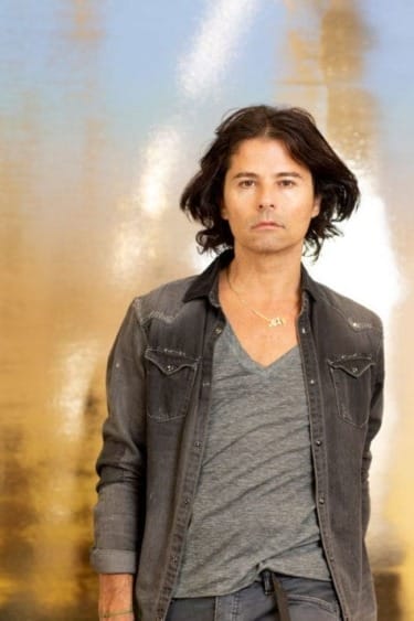 Person with long dark hair wearing a grey shirt and black jacket standing in front of a blurred golden background.