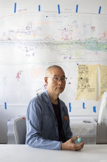 Person sitting at a desk in front of a large whiteboard with drawings and diagrams, holding a small object.