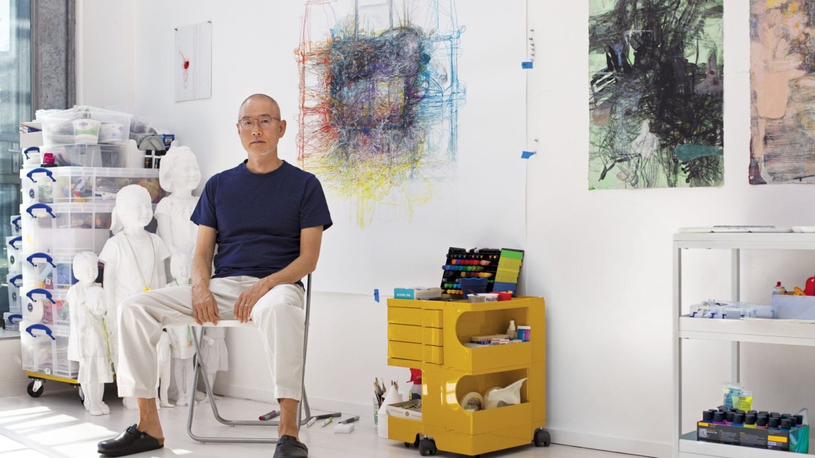 Man sitting in a bright art studio with colorful artworks on the walls and art supplies on shelves and a cart.
