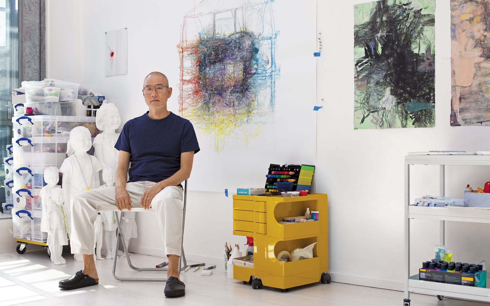 Man sitting in a bright art studio with colorful artworks on the walls and art supplies on shelves and a cart.