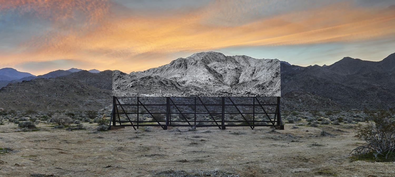 Outdoor art installation merging a desert mountain scene with a photo panel, under a colorful sunset sky.