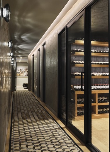Elegant hallway with patterned rug leading to a glass-enclosed wine cellar featuring wooden shelves stocked with wine bottles.