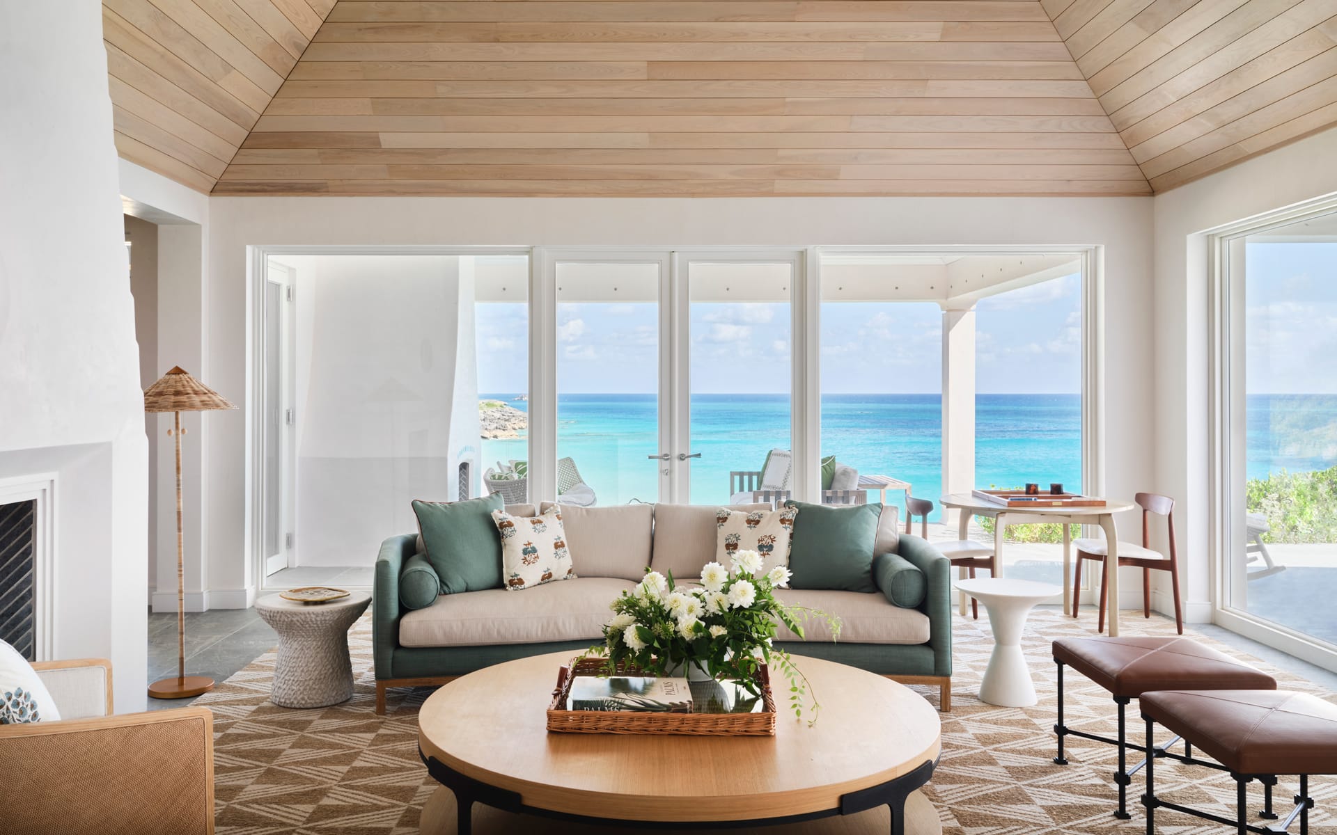 Spacious living room with sea view, wooden ceiling, beige sofa, and round table with flowers, overlooking a sunny beach.