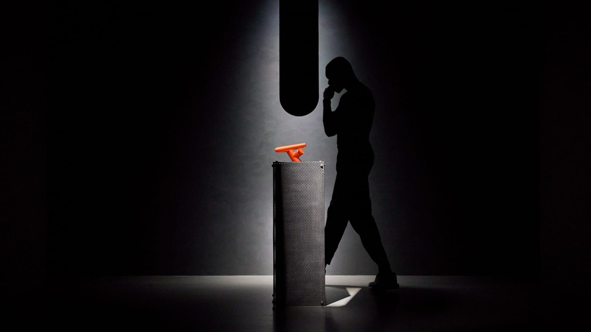 Silhouette of a person pondering near a pedestal with a bright orange object in a dark, minimalist setting.