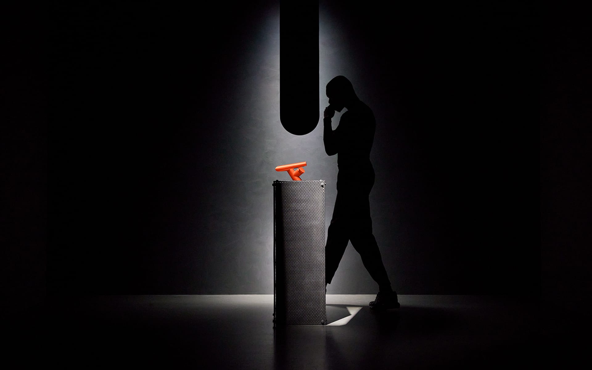 Silhouette of a person pondering near a pedestal with a bright orange object in a dark, minimalist setting.
