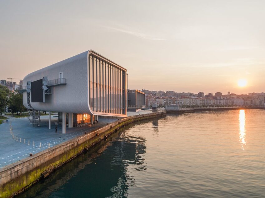 Modern waterfront building with large windows at sunset, reflecting on calm water, surrounded by city skyline.