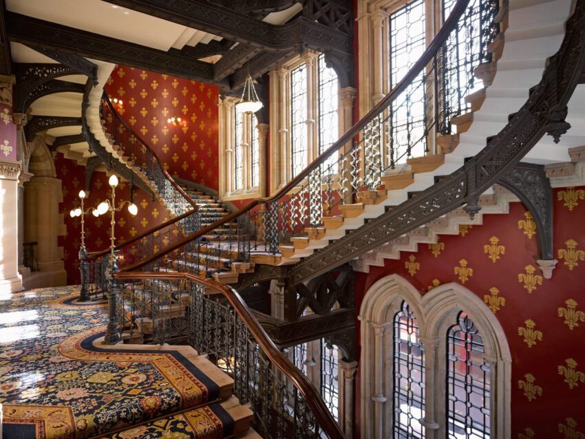 Grand staircase with ornate railing, patterned carpet, and red walls in a historic building interior.