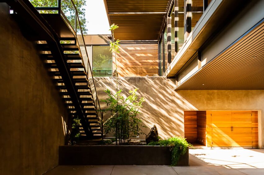 Modern architectural courtyard with sunlight filtering through, featuring a staircase, plants, and large wooden doors.