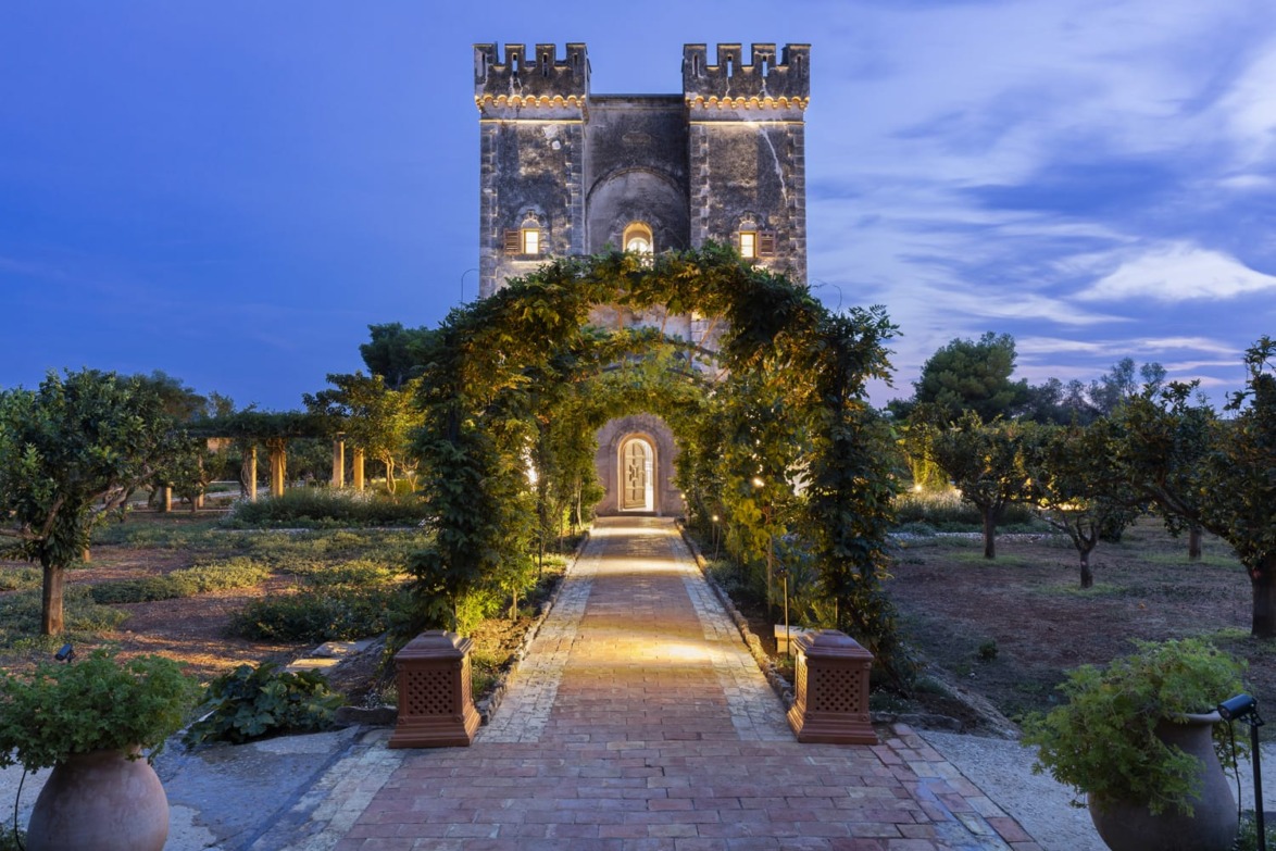 Historic stone tower with illuminated pathway surrounded by lush gardens and trees during twilight, featuring a clear sky.