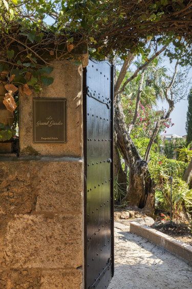Open wrought iron gate leading to a lush garden with trees and flowers under a clear blue sky.