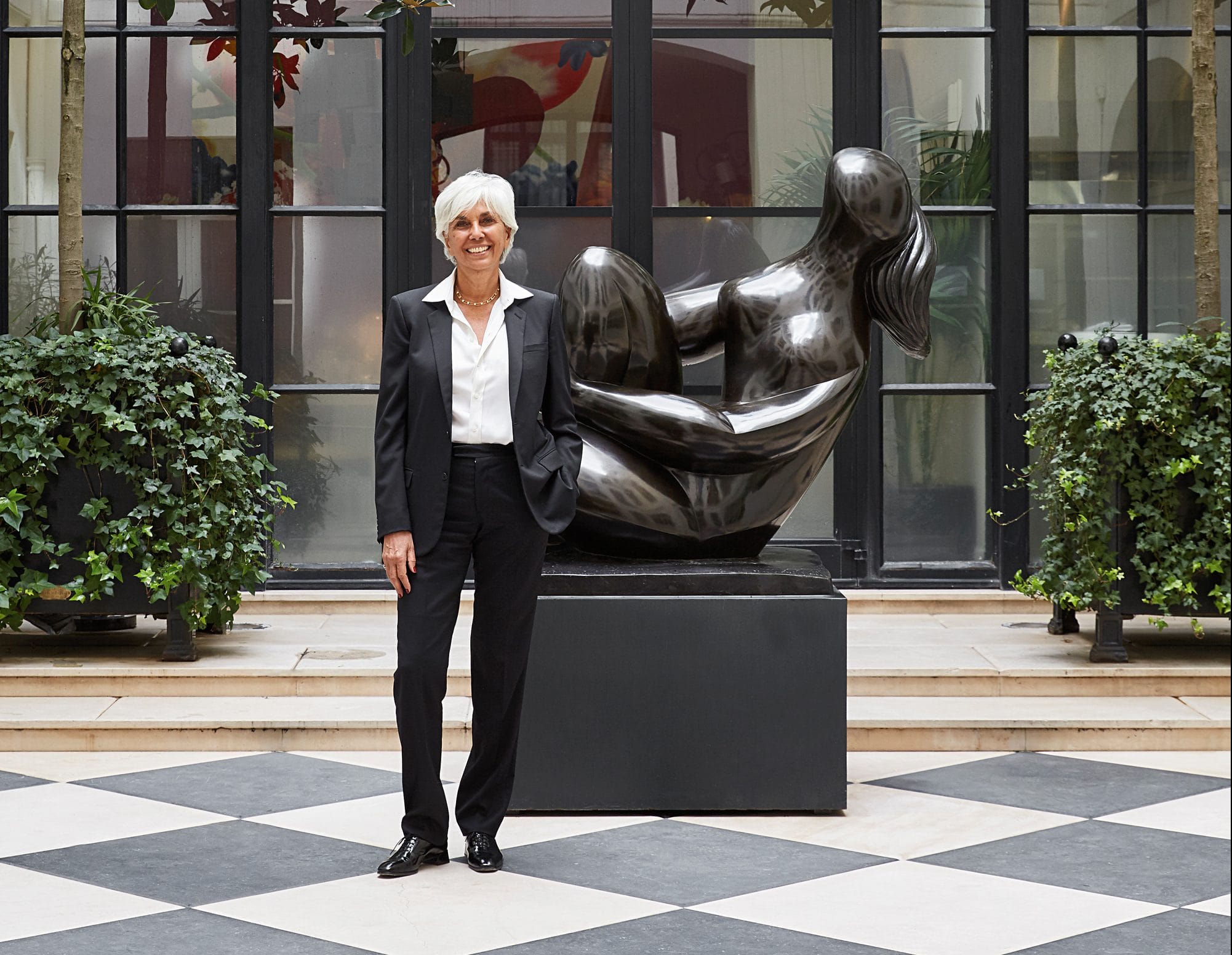 Person in a suit standing in a stylish atrium with a modern sculpture in the background and plants on either side.