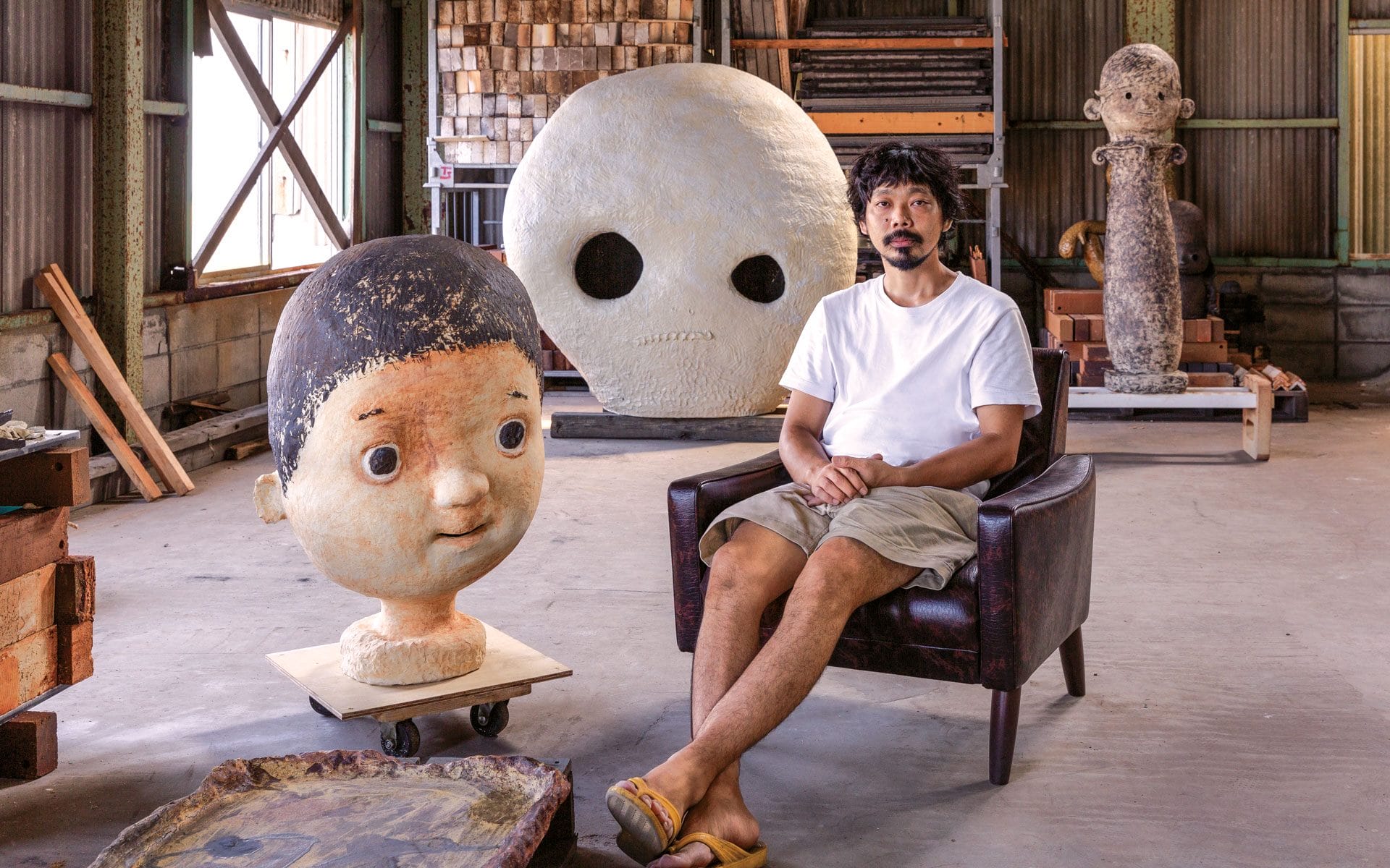 Person sitting in a chair in an art studio with large sculptures of stylized human heads in the background.