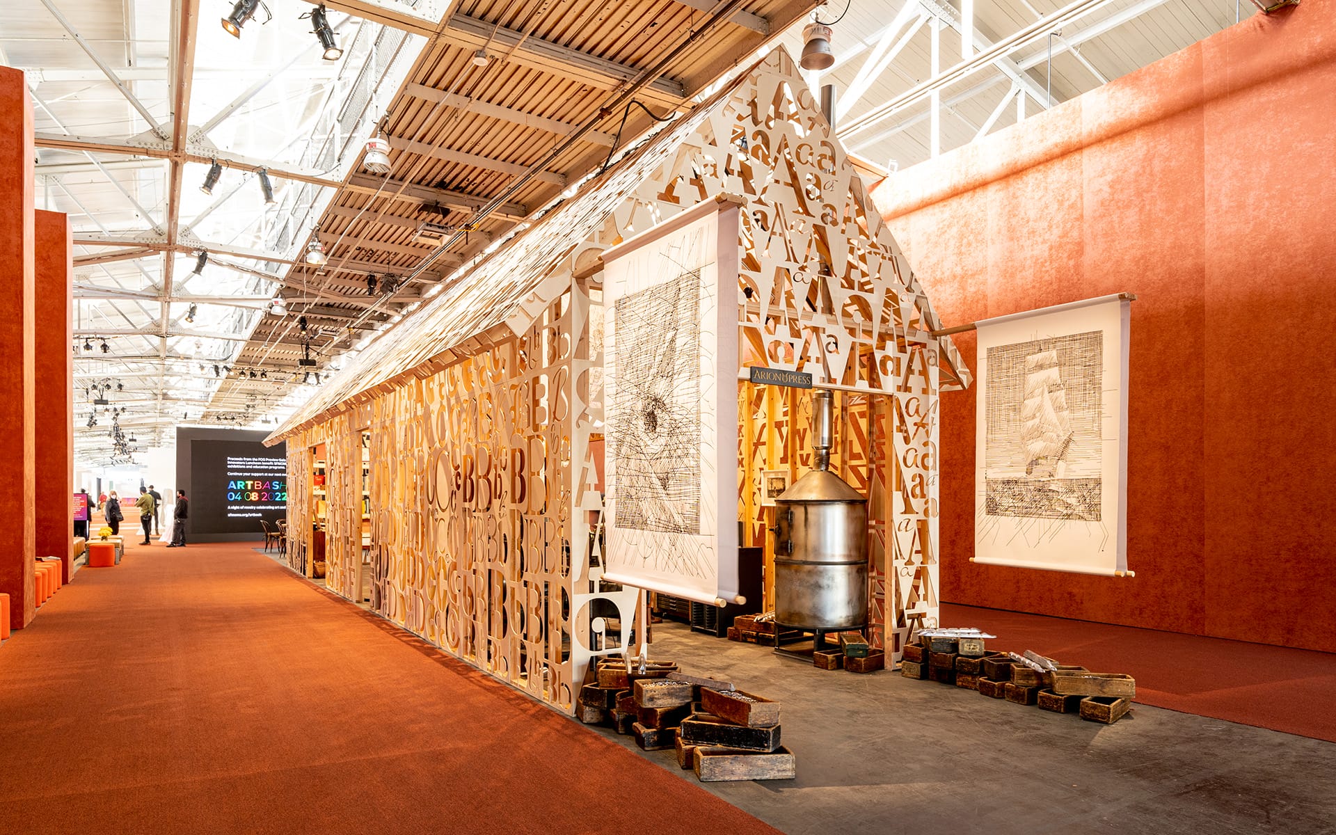 Art exhibit with a geometric structure featuring alphabet cutouts, orange walls, and framed artworks, inside a large, well-lit space.