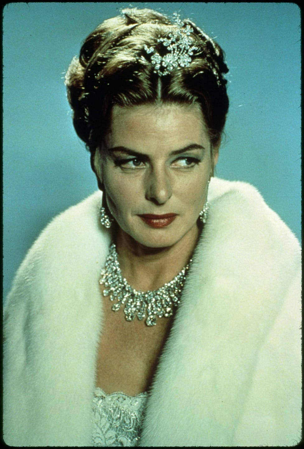 Woman wearing a white fur coat, diamond tiara, and jewelry against a blue background, looking off to the side