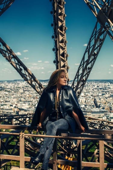 Person sitting on Eiffel Tower structure, wearing a stylish black jacket, with Paris cityscape in the background.