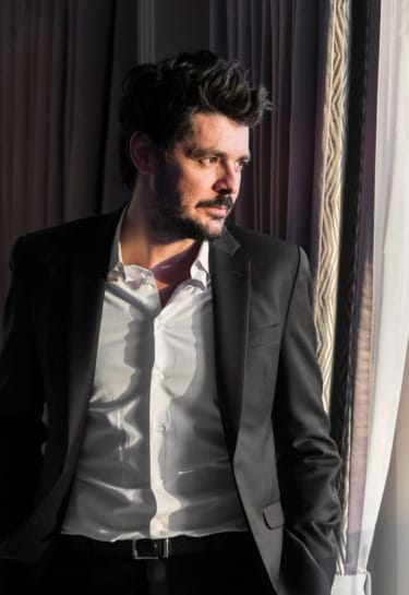 Man in a suit standing near a window, looking thoughtfully outside.