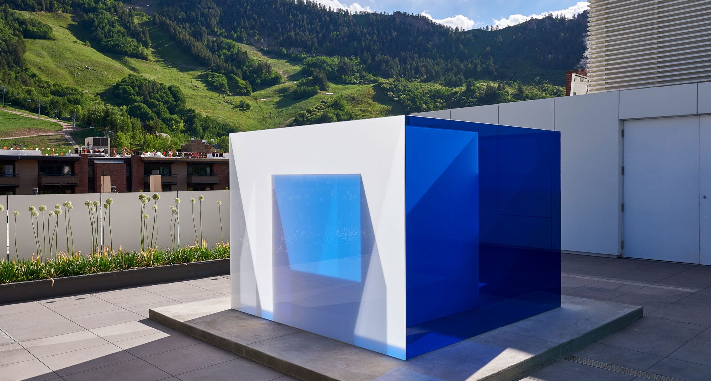 Outdoor art installation with blue and white geometric glass structure on a rooftop, surrounded by greenery and mountains.