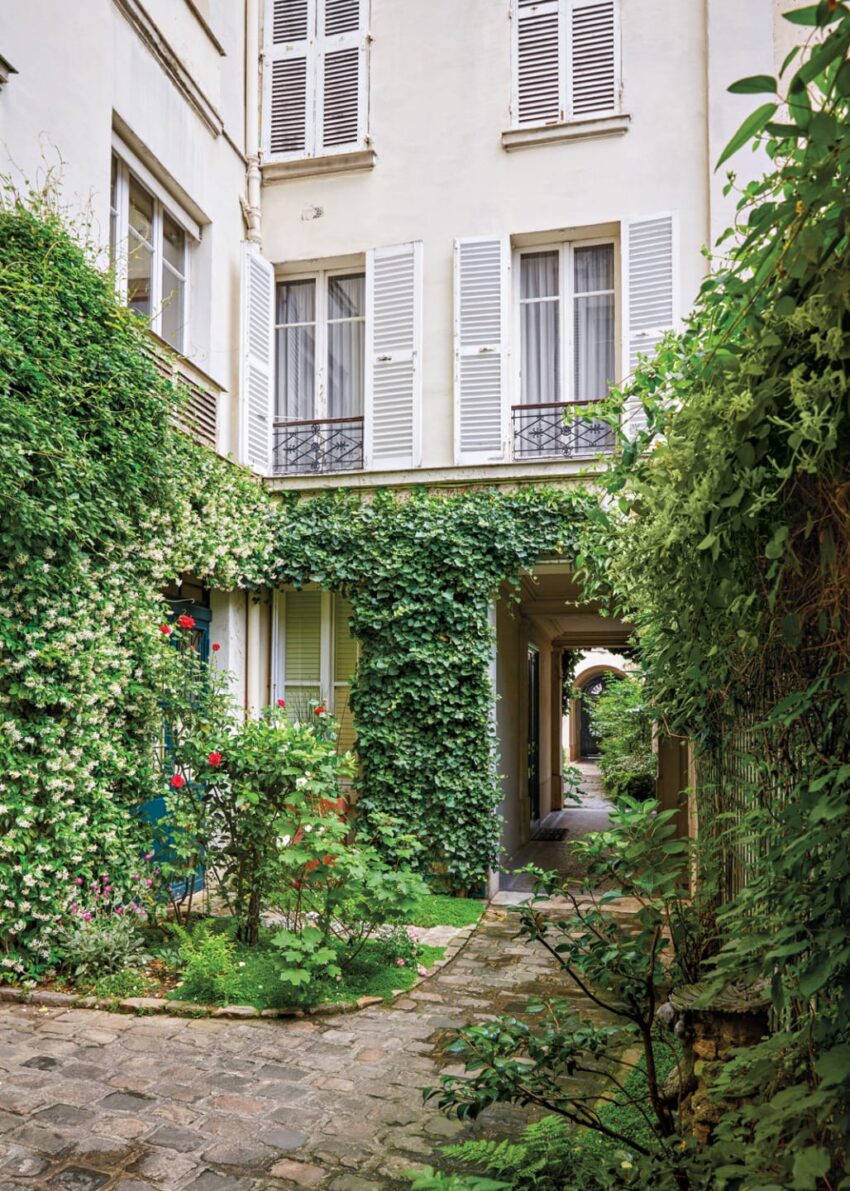 Courtyard with green ivy-covered buildings, flowering plants, and a stone path leading to an arched passageway.