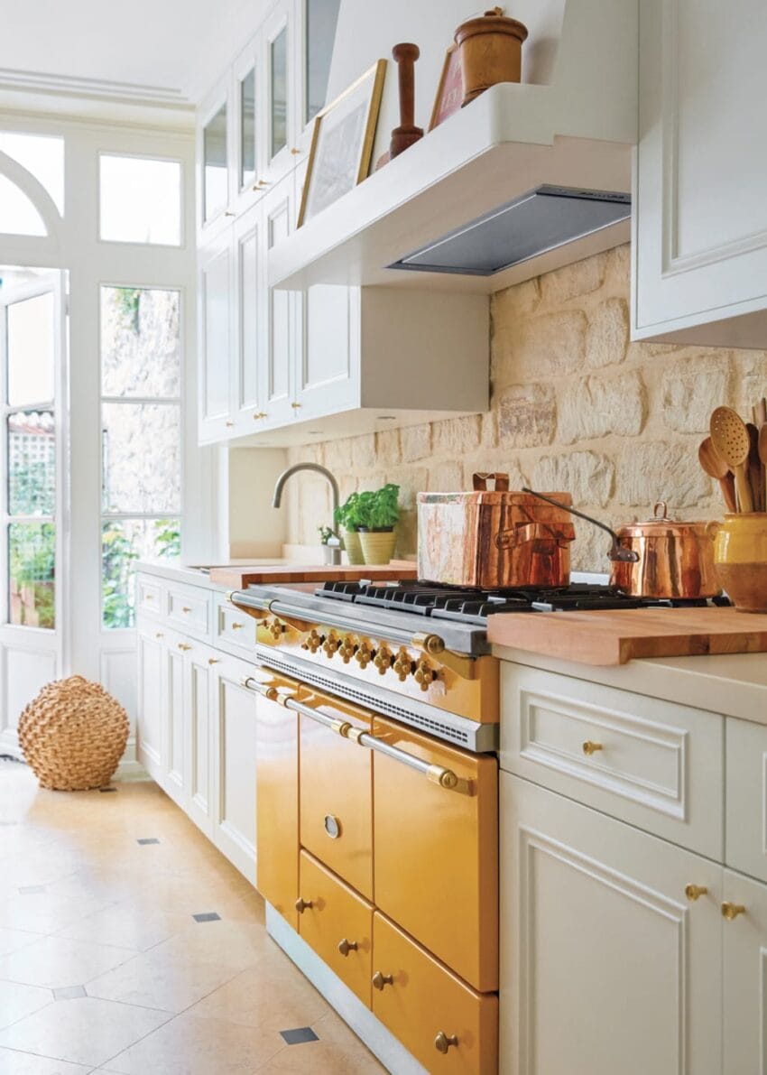 Modern kitchen with white cabinets, brass knobs, yellow oven, copper cookware, and brick backsplash under natural light.
