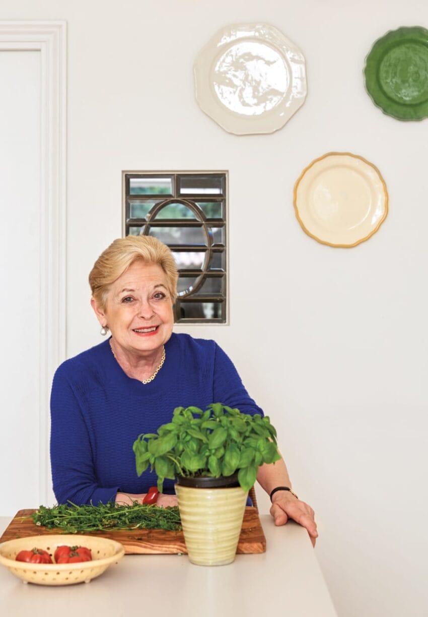 Woman in blue sweater with herbs and potted plant in kitchen, decorative plates on wall in background