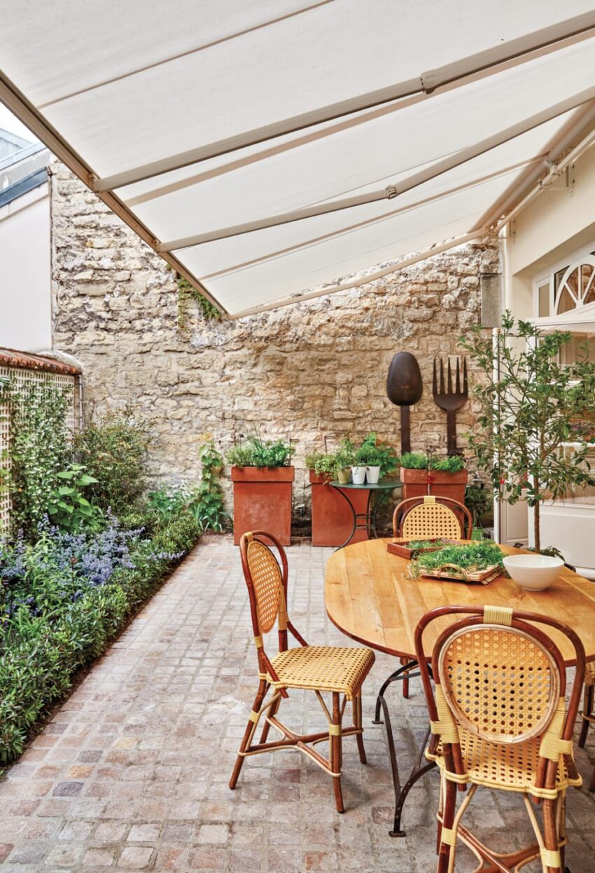 Charming outdoor patio with rattan chairs, a wooden table, potted plants, and a stone wall backdrop under a white awning.