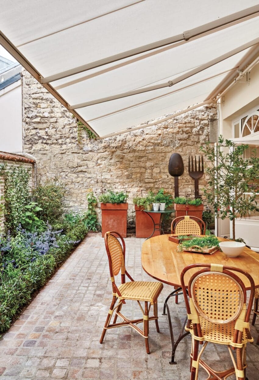 Charming outdoor patio with rattan chairs, a wooden table, potted plants, and a stone wall backdrop under a white awning.